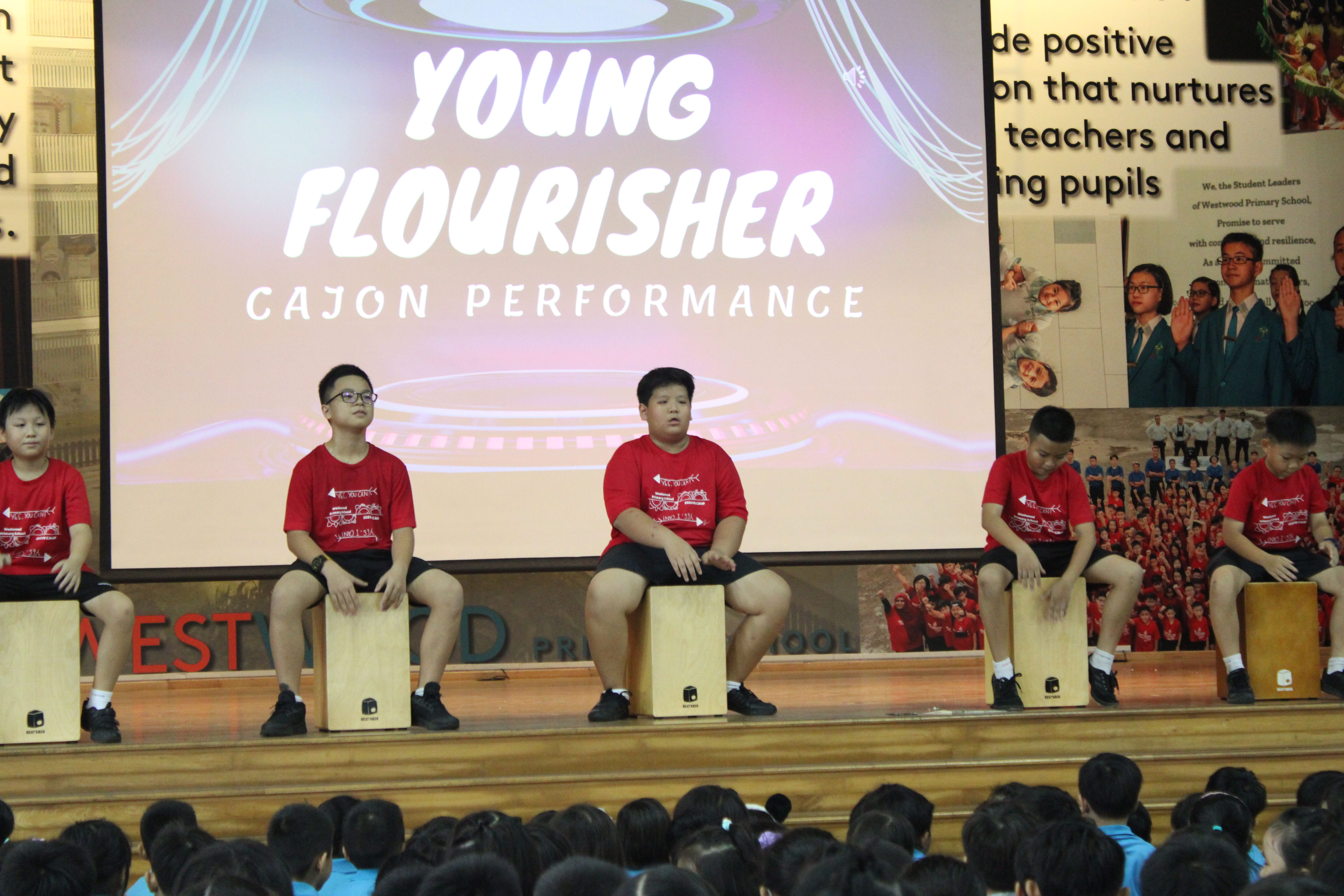Five children in red shirts sit on cajon drums on a school stage performing for an audience below.